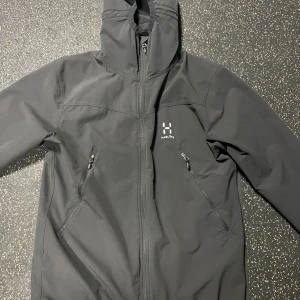 Svart vindjacka från Haglöfs - Svart vindjacka från Haglöfs med huva och dragkedja framtill. Jackan har två snedställda fickor med dragkedja och diskret logga på bröstet. Perfekt för blåsiga dagar och outdoor-aktiviteter. Använd två gånger.