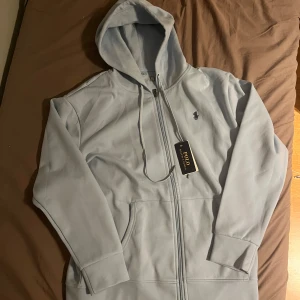 Ljusblå hoodie från Polo Ralph Lauren - Snygg ljusblå hoodie från Polo Ralph Lauren med dragkedja och huva. Klassisk logga broderad på bröstet och praktiska fickor framtill. Perfekt för en chill och stilren look. Tillverkad i mjuk bomull för extra komfort.