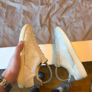 Gucci ace sneakers i skinn - Snygga vita sneakers från Gucci med perforerad logga på sidan och grå hälparti med Gucci-tryck. Skorna har rund tå, platt sula och klassisk snörning. Tillverkade i mjukt skinn med detaljerad sula och stilren design som passar till jeans eller shorts.