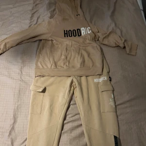 Hoodrich dress  - Säljer en snygg beige dress från Hoodrich med stor logga i svart och vitt på bröstet. Tröjan har huva med snörning och en stor magficka. Perfekt för dig som gillar streetwear och vill ha en chill outfit. STORLEK M på hoodie, STORLEK L på byxorna passar även de som har M eftersom de är små i storleken. 