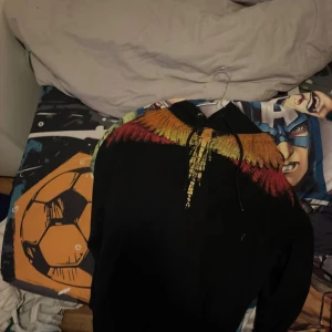 Marcelo Burlon svart hoodie med färgglada vingar - Säljer en svart hoodie från Marcelo Burlon med ett fett tryck av färgglada vingar i rött, orange och gult över axlarna och bröstet. Hoodien har huva med snörning och lång ärm, perfekt för dig som vill sticka ut med en unik design.