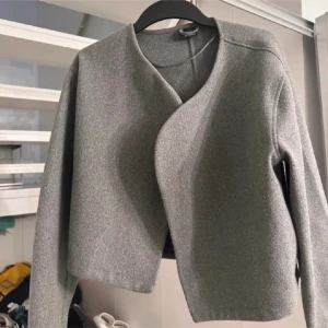 Grå omlott cardigan jacka - Snygg grå cardigan jacka med omlott-design och lång ärm. Jackan har en minimalistisk look utan synliga knappar eller dragkedja och är lätt croppad. Perfekt för dig som gillar stilrena och moderna plagg. lite mer sliten än bilden visar