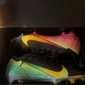 Nike fotbollsskor med regnbågsfärg - Säljer ett par Nike fotbollsskor med svart ovandel och stora gula swoosh-loggor. Skorna har en färgskiftande sula och överdel i regnbågsfärger med textdetaljer i vitt. Ovandelen är i syntetmaterial och har snörning för bra passform. Perfekta för dig som vill sticka ut på planen. Det har släppt lite på vänster skon men går fortfarande utmärkt att spela med dom, Se bild 3