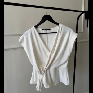 Vit omlottopp från Zara - Säljer en stilren vit omlottopp från Zara med v-ringning och knyt i sidan. Toppen är ärmlös och har en lätt utsvängd passform som ger en snygg siluett. Perfekt att matcha med jeans eller kjol för en fräsch look. Den har dragkedja på sidan