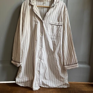 Randig vit skjorta med bruna detaljer - Vit skjorta med tunna bruna och vinröda ränder, klassisk krage och bröstficka. Skjortan har långa ärmar och knäppning framtill. Materialet känns mjukt och luftigt, perfekt för en chill look.