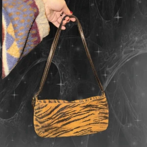 Tiger-mönstrad axelväska från Vera Pelle - Snygg axelväska i tiger-mönstrat läder från Vera Pelle. Väskan har en brun justerbar axelrem med spänne och dragkedja upptill. Insidan är svart med en liten innerficka med dragkedja. Perfekt storlek för det viktigaste.