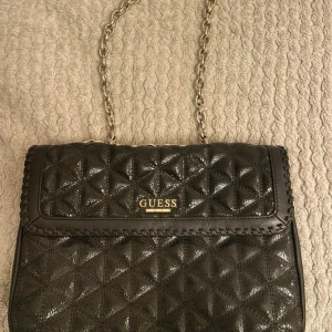Svart quiltad axelväska från Guess - Snygg svart axelväska från Guess med quiltad yta i geometriskt mönster. Väskan har en kedja i guldfärg som axelrem och ett stort fack med dragkedja inuti. Guess-loggan i metall framtill ger en lyxig touch. Perfekt storlek för det viktigaste.
