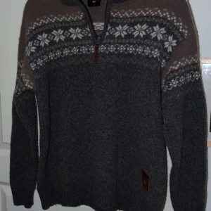 Mörkgrå stickad tröja med mönster - Mysig mörkgrå stickad tröja med klassiskt vitt och grått nordiskt mönster över bröstet och axlarna. Tröjan har lång ärm och en praktisk half zip-dragkedja vid halsen. Perfekt för kyliga dagar och snygg till jeans.