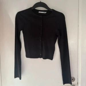 Svart croppad topp med knappar - Svart croppad topp från lager 157 i storlek XS. Modellen har långa ärmar, rund hals och knappar hela vägen framtill. Materialet är ribbad bomull som ger en skön och stretchig känsla. Perfekt att styla med jeans eller kjol för en clean look.
