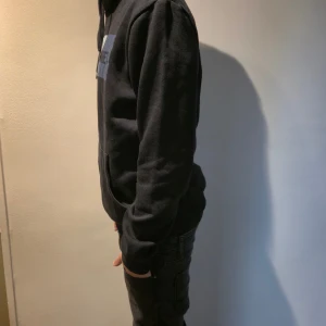 Svart hoodie från Jack & Jones - Svart hoodie från Jack & Jones med stor logga i rött, vitt och blått framtill. Tröjan har huva med snörning och en stor magficka. Mjuk och skön, perfekt för chill dagar. Långärmad och i klassisk passform. Hör av er om ni har funderingar. Jag själv är 165-170 på bilden.