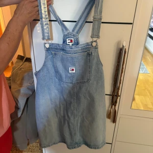 Blå jeansklänning från Tommy Hilfiger - Säljer en snygg blå jeansklänning från Tommy Hilfiger med klassiska hängslen och en stor ficka framtill. Klänningen har logotypen på både fickan och bröstet, och är perfekt för dig som gillar en avslappnad och cool stil.
