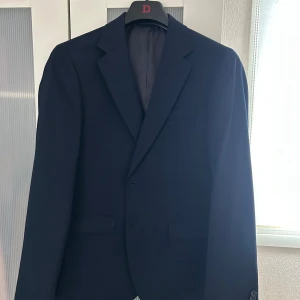 Mörkblå kostym Dressmann slim fit - Säljer en stilren mörkblå kostym från Dressmann i storlek 50 Regular, slim fit. Använd en gång. Kavajen har klassisk krage, två fickor med lock och knäppning framtill. Byxorna är raka och har hällor för bälte. Byxorna är i storlek 33/30. Ordinarie pris: 2 600kr. Pris kan diskuteras vid snabb affär.