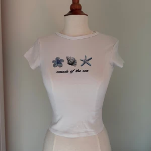 Vit croppad topp med tryck - Säljer en vit croppad topp med korta ärmar och tryck framtill med blommor, snäcka och sjöstjärna samt texten 'sounds of the sea'. T-shirten har en tight passform och är gjord i mjuk bomull. Perfekt för dig som gillar strandvibbar och enkel stil.