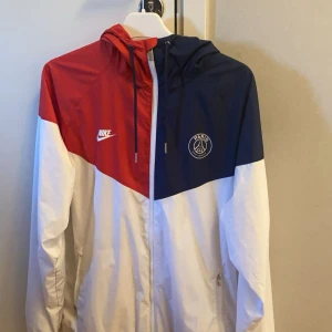 Nike PSG vindjacka röd/blå/vit - Snygg PSG vindjacka från Nike i rött, blått och vitt. Jackan har huva med snörning, dragkedja framtill och två fickor med dragkedja. PSG-logga och Nike Swoosh på bröstet. Perfekt för dig som vill ha sportig stil och visa vilket lag du supportar.