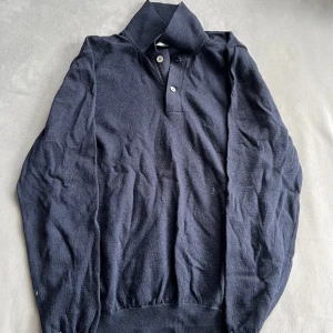 Mörkblå stickad pikétröja - Mörkblå stickad pikétröja med klassisk krage och två knappar framtill. Tröjan har långa ärmar och ribbade muddar vid ärmslut och nederkant. Perfekt för dig som gillar en stilren och enkel look.