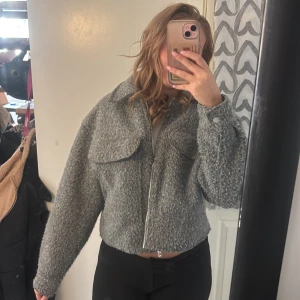 Grå teddyjacka med dragkedja - Säljer en grå teddyjacka med croppad passform och stora bröstfickor. Jackan har en rejäl krage, lång ärm och stängs med dragkedja framtill. Perfekt för dig som gillar en chill och trendig look.