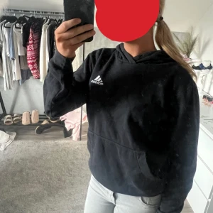 Svart hoodie från Adidas - Svart hoodie från Adidas med vit logga på bröstet. Klassisk modell med huva och känguruficka framtill. Perfekt för en avslappnad och sportig stil. Mjuk och skön, passar till jeans eller träningsbyxor.
