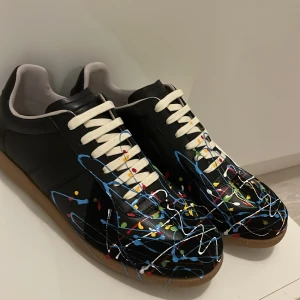 Maison Margiela  - Maison Margiela sneakers i svart skinn med coola färgstänk i blått, vitt, rött, gult och grönt. Skorna har vit snörning och en klassisk gummisula i beige. Snygg och unik design som sticker ut, perfekt för dig som gillar stil. Helt nya med box aldrig använd storlek 44 köpte de på Plick för 2200. jag ska sälja dem för 1499 kan sänka pris vid snabb affär