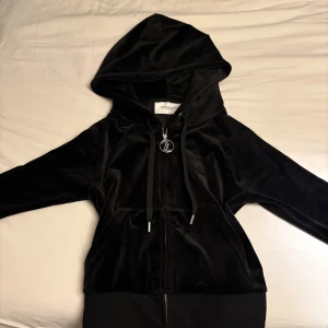Svart velour hoodie från Juicy Couture - Säljer en svart hoodie från Juicy Couture i mjuk velour med huva och dragkedja. Tröjan har långa ärmar, snörning vid huvan och en stor ficka framtill. Ikonisk JC-logga på bröstet och snygg metall-detalj på dragkedjan.