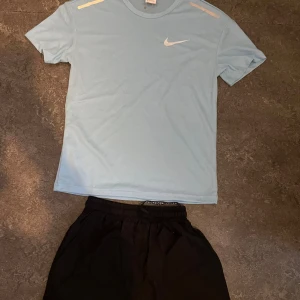 Ljusblå Nike t-shirt med logga - Fräsch ljusblå t-shirt från Nike med klassisk vit swoosh på bröstet och diskreta vita detaljer på axlarna. T-shirten har rund hals och korta ärmar, perfekt för dig som gillar sportig och clean stil. Materialet är lätt och andas, vilket gör den grym för träning. 