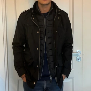 J.Lindeberg field jacket - Svart field jacket från J.Lindeberg med flera fickor, axelklaffar och quiltad innerdel. Jackan har dragkedja och knappar framtill samt hög krage. Det är en inbyggd väst innanför och perfekt för höst och vinter, snygg och stilren design som funkar till jeans och sneakers. Pris kan diskuteras💯