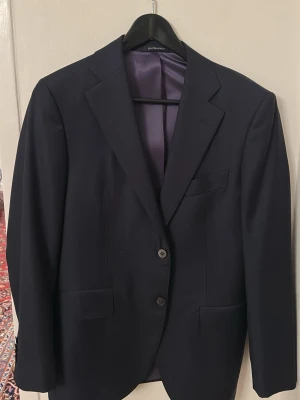 Mörkblå kavaj från SuitSupply Lazio - Stilren svart kavaj från SuitSupply, modell Lazio. Slim fit med enkelknäppning och två knappar framtill. Tillverkad i 100% ull med slät finish och klassiska fickor. Perfekt för en dressad look.