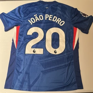 Chelsea João Pedro #20 Nike matchtröja - Chelsea FCs hemmatröja från Nike med João Pedro och nummer 20 på ryggen. Tröjan är blå med vita detaljer, har korta ärmar och Premier League-logga på ärmen. Tillverkad i lätt och ventilerande funktionsmaterial för maximal komfort.