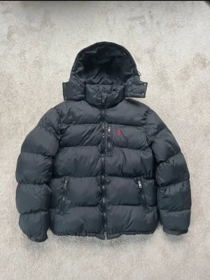 Svart dunjacka från Polo Ralph Lauren - Säljer en svart dunjacka från Polo Ralph Lauren med avtagbar huva och dragkedja framtill. Jackan har två sidofickor med dragkedja och en liten röd logga på bröstet. Perfekt för kalla vinterdagar och riktigt skön att bära.