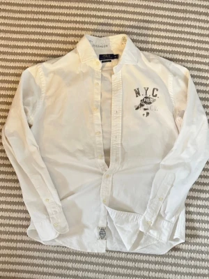 Ralph Lauren N.Y.C skjorta - Vit skjorta från Polo Ralph Lauren med tryck på bröstet där det står N.Y.C. och POLO. Skjortan har klassisk krage, långa ärmar och knäppning framtill. Perfekt för dig som gillar en clean och sportig stil.