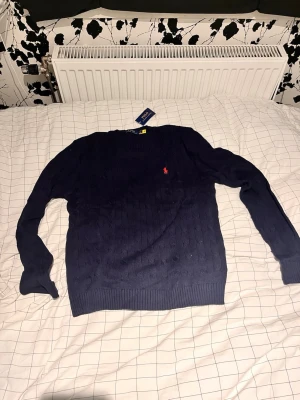 Mörkblå stickad tröja Polo Ralph Lauren - Mörkblå stickad tröja från Polo Ralph Lauren med rund halsringning och klassisk röd logga broderad på bröstet. Tröjan har ribbstickade muddar och är tillverkad i mjukt material. Perfekt för en stilren och avslappnad look.