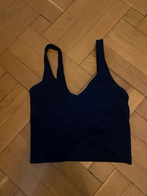 Mörkblå sport-bh från Lululemon - Snygg mörkblå sport-bh från Lululemon med breda axelband och V-ringning. Tillverkad i ett stretchigt och mjukt material som passar perfekt för träning eller yoga. Diskret Lululemon-logga på ryggen. Enkel och stilren design.