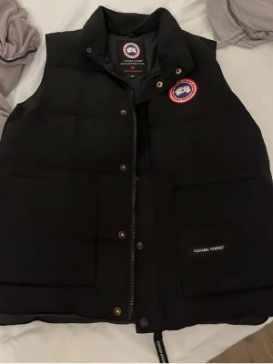 Svart dunväst från Canada Goose - Snygg svart dunväst från Canada Goose med klassisk logga på bröstet och två stora fickor framtill. Västen har dragkedja och tryckknappar samt är fodrad med dun för extra värme. Perfekt för lager-på-lager och streetstyle.
