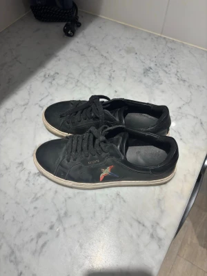 Svarta Axel Arigato  - Snygga svarta sneakers från Axel Arigato med klassisk låg profil och vit sula. Skorna har en färgglad broderad fågel på sidan och guldfärgad logga. Tillverkade i läder med svarta skosnören och stilren design som passar till det mesta.