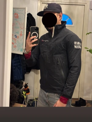 Svart vindjacka från Sail Racing - Snygg svart vindjacka från Sail Racing med dragkedja framtill, hög krage och logga på bröstet samt ärmen. Jackan har två fickor med dragkedja och en sportig, stilren look. Perfekt för blåsiga dagar och enkel att matcha med jeans.