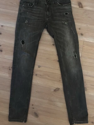 Svarta slitna jeans från Icono Couture - Säljer ett par svarta jeans från Icono Couture med flera slitningar och hål på benen för en riktigt edgy look. Jeansen har klassisk femficksdesign och raka ben. Perfekta för dig som gillar streetstyle och vill sticka ut.