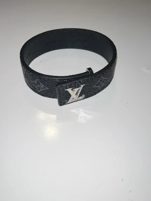 Louis Vuitton svart läderarmband - Stilrent svart armband i präglat läder från Louis Vuitton med ikoniskt LV-spänne i silverfärgad metall. Armbandet har en modern och minimalistisk design som passar perfekt till din outfit. Snygg accessoar för dig som gillar exklusiva detaljer.