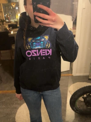 Svart Kenzo hoodie med tigertryck - Svart hoodie från Kenzo med ikoniskt tigertryck i blått, gult och lila samt texten 'KENZO PARIS' i lila framtill. Klassisk känguruficka och huva med dragsko. Perfekt för dig som gillar streetwear och vill sticka ut med en färgstark detalj.