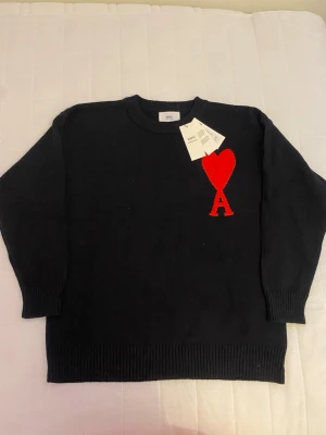 Svart stickad Ami Paris tröja - Säljer en svart stickad tröja från Ami Paris med deras ikoniska röda hjärta och bokstaven A broderat på bröstet. Tröjan har rund halsringning, ribbade muddar och är gjord i mjukt material som passar perfekt till höst och vinter. Ärmläng: 57cm Läng: 66cm