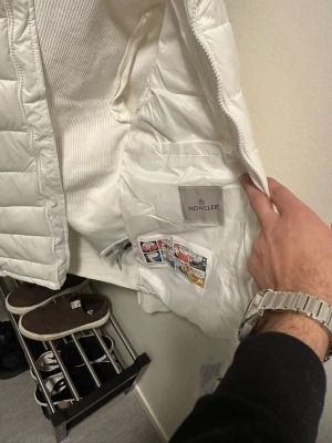 Vit dunväst från Moncler - Snygg vit dunväst från Moncler med quiltad design och dragkedja. Västen har en klassisk look och är fodrad med mjukt material. Perfekt att slänga över en hoodie eller tröja för extra värme. Märkeslogga på insidan och coola detaljer.