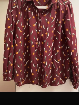 Blommig vinröd skjorta med knappar - Snygg vinröd skjorta med blommigt mönster i gult och grönt. Skjortan har långa ärmar, klassisk krage och knäppning framtill. Perfekt för dig som vill ha en färgglad och unik look. En knapp är bruten och det är ett hål som synts ihop på ena armen längs sömnen. 