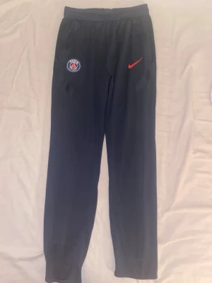 Mörkblå PSG sportbyxor från Nike - Säljer ett par svarta mjukisbyxor från Nike med PSG-logga på vänster sida och röd Nike swoosh på höger. Byxorna har resår i midjan och vid bensluten, samt fickor på sidorna. Perfekta för chill eller träning. Jätte bra skick men håller på rensa min garderob.