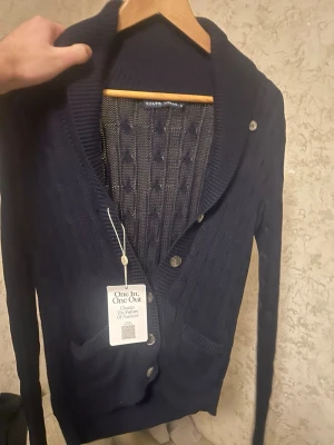 Mörkblå kabelstickad kofta Ralph Lauren - Snygg mörkblå kabelstickad kofta från Ralph Lauren med sjalkrage och knappar framtill. Koftan har två fickor och ribbade muddar. Perfekt för lager på lager och ger en klassisk look.