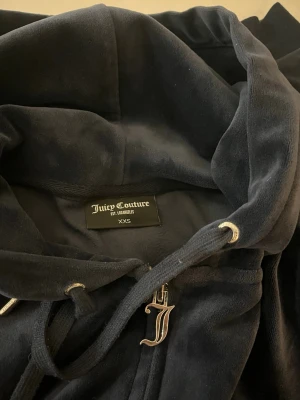 Svart velour hoodie Juicy Couture - Mjuk och lyxig svart velour hoodie från Juicy Couture med dragkedja framtill, snörning i huvan och metalliska detaljer. Klassisk Juicy-logga broderad på bröstet och ett snyggt J-hänge på dragkedjan. Perfekt för en avslappnad och trendig look.