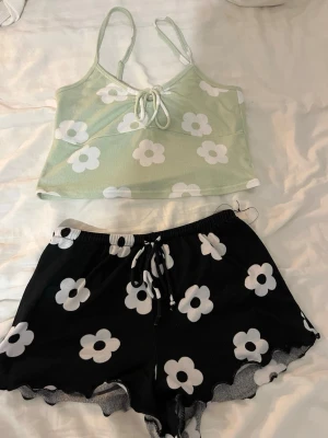 Blommig pyjamas med linne och shorts - Söt pyjamas  med ett ljusgrönt linne med smala axelband och knyt framtill samt svarta shorts med vita blommor och dragsko i midjan. Båda delar har ett väldigt mjukt och behagligt klimat material. Köpt på new yorker i storlek S💚