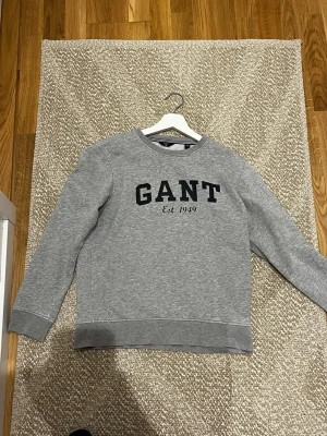 Grå sweatshirt från GANT - Klassisk grå sweatshirt från GANT med svart tryck på bröstet där det står 'GANT Est. 1949'. Tröjan har rund halsringning, långa ärmar och ribbade muddar vid ärmslut och nederkant. Perfekt för en avslappnad och stilren look. Storleken är 170 cm