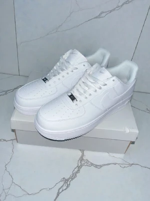 Nike Air Force 1 vita sneakers - Klassiska Nike Air Force 1 sneakers i helvitt läder med perforerad tå, vit sula och diskret Nike-logga på sidan. Skorna har lågt skaft och vita skosnören. Perfekt för dig som gillar en clean och tidlös stil.