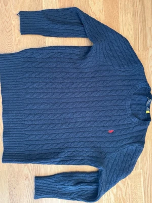 Mörkblå kabelstickad tröja Polo Ralph Lauren - Mörkblå kabelstickad tröja från Polo Ralph Lauren med rund halsringning och röd broderad logga på bröstet. Tröjan har långa ärmar och ribbade muddar. Perfekt för en klassisk och stilren look.