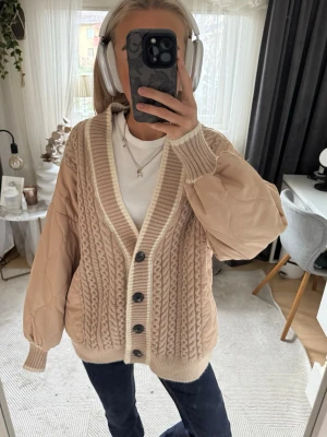 Beige kabelstickad cardigan jacka - Trendig beige cardigan jacka med kabelstickat mönster framtill och släta, quiltade ärmar. Djup v-ringning, bred ribbad kant och stora svarta knappar. Passar till allt!!🫶🏼