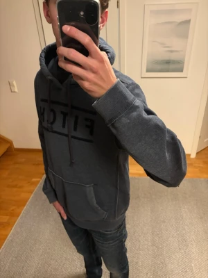 Grå/blå hoodie från Fitch -  Grå/blå hoodie med stor magficka och dragsko i huvan. Nån små fläck som går bort i tvätten.Tjockt och mjukt material, perfekt för chill dagar. Stor logga med texten 'FITCH' framtill. Snygga sömdetaljer och klassisk passform. Storlek M kan passa L.