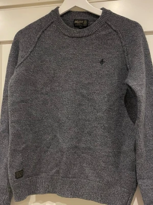 Grå stickad tröja från Morris - Snygg gråmelerad stickad tröja från Morris med rund halsringning och diskret broderad logga på bröstet. Tröjan har ribbstickade muddar vid ärmslut och nederkant. Perfekt för lager-på-lager och en stilren look.